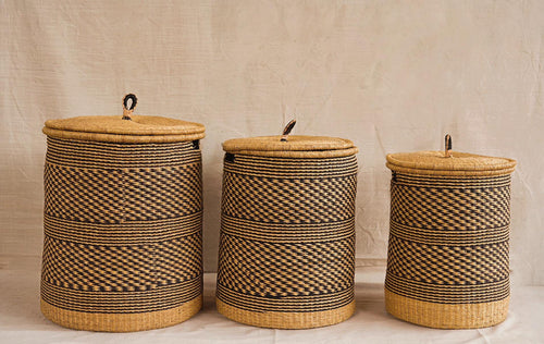 Three cylindrical Bolga storage baskets with lids, in different sizes, featuring a natural weave pattern and leather-wrapped handles.